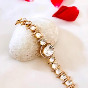 May include: A gold bracelet featuring clear, oval gemstones and a large, square-cut central stone. The bracelet is displayed on a white stone, with red rose petals in the background. This jewellery is suitable for special events.