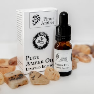 May include: A white box with black text that reads "Pinus Amber Pure Amber Oil Limited Edition". A small amber glass bottle with a black dropper and a white label that reads "Pinus Amber Amber Oil Limited Edition" is in front of the box. The bottle is surrounded by small amber stones.
