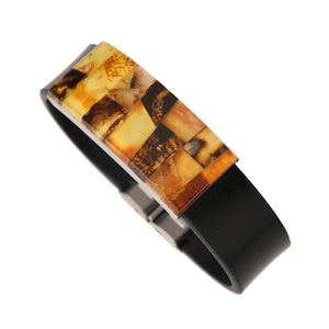 Puede incluir: Un brazalete de cuero negro con una incrustación rectangular de ámbar. El ámbar es un mosaico de diferentes tonos de amarillo y marrón.