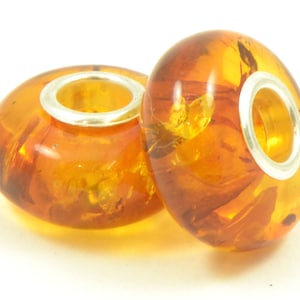 Op de afbeelding: Twee amber kralen met zilveren accenten. De kralen zijn rond en hebben een doorschijnende uitstraling. De amber heeft een warme honingkleur en bevat kleine insluitsels.