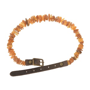 Puede incluir: Collar con piedras irregulares de color ámbar. Se adjunta a una correa de cuero marrón oscuro con una hebilla de metal y un cierre de botón. Las piedras de ámbar varían en tamaño y tono.