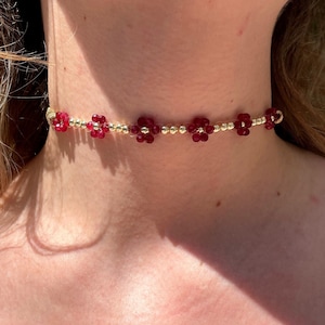 Può includere: Una collana girocollo dorata con piccole perline dorate e perline rosse a forma di fiore. La collana, indossata attorno al collo, mette in risalto il delicato design floreale e il contrasto tra l'oro e il rosso. Lunghezza di circa 35-40 cm.