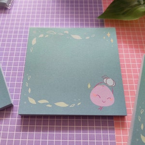 May include: Square teal sticky notes with a cute cartoon character illustration. The character is a pink fruit with a smiling face and a small hat. The notes have a border of leaves and stars.