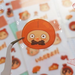 Może przedstawiać: Przezroczysta naklejka przedstawiająca postać z kreskówki w pomarańczowym stroju z brązowymi wąsami, trzymana pęsetą. Arkusz naklejek zawiera inne obrazy z kreskówek i japoński tekst w kolorze pomarańczowym.