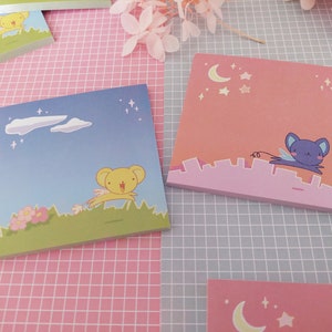 Könnte beinhalten: Quadratische Haftnotizen mit Cartoon-Illustrationen. Eines zeigt einen gelben, bärenartigen Charakter, der über ein grünes Feld unter blauem Himmel mit Wolken springt. Ein anderes zeigt einen lila, bärenartigen Charakter, der über einer rosa Stadtlandschaft fliegt.