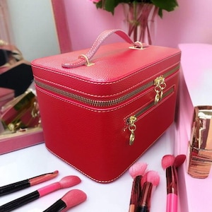 Puede incluir: Un estuche de maquillaje rojo con asa y cremalleras doradas. El estuche es rectangular y se encuentra sobre una superficie blanca con brochas de maquillaje. Un ramo de rosas rosas está en el fondo.