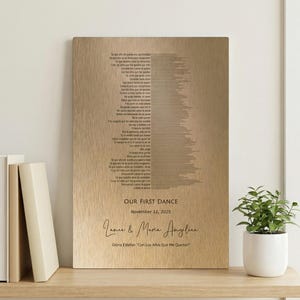 May include: A wooden plaque with engraved text. The text reads "OUR FIRST DANCE" and "November 12, 2025" along with names and a song title. The plaque is displayed on a wooden shelf with books and a potted plant.