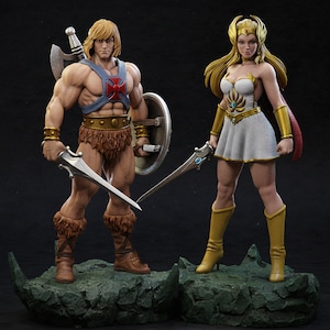 以下が含まれることがあります： 岩場に立つ、He-ManとShe-Raの精巧なフィギュア。He-Manは筋肉質で、剣と盾を持ち、ハーネスと毛皮のブリーフを着用。She-Raは白いドレス、金のアクセント、ブーツを身に着け、剣も持っています。フィギュアは暗い背景に配置されています。