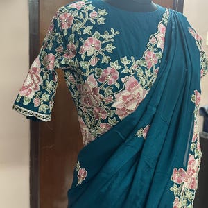 Puede incluir: Sari de color turquesa con intrincado bordado floral en rosa y verde. El sari presenta un corpiño y mangas adornados, mostrando una mezcla de diseño tradicional y elegancia moderna. La tela se drapea con gracia, resaltando los patrones florales.
