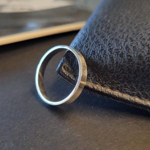 May include: A silver ring with a smooth, polished finish. The ring is lying on a black leather surface.