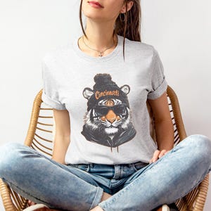 Puede incluir: Una camiseta gris con un gr&aacute;fico de un tigre que lleva una boina negra con la palabra "Cincinnati" en ella. El tigre lleva gafas de sol y tiene un patr&oacute;n de rayas blancas y negras.
