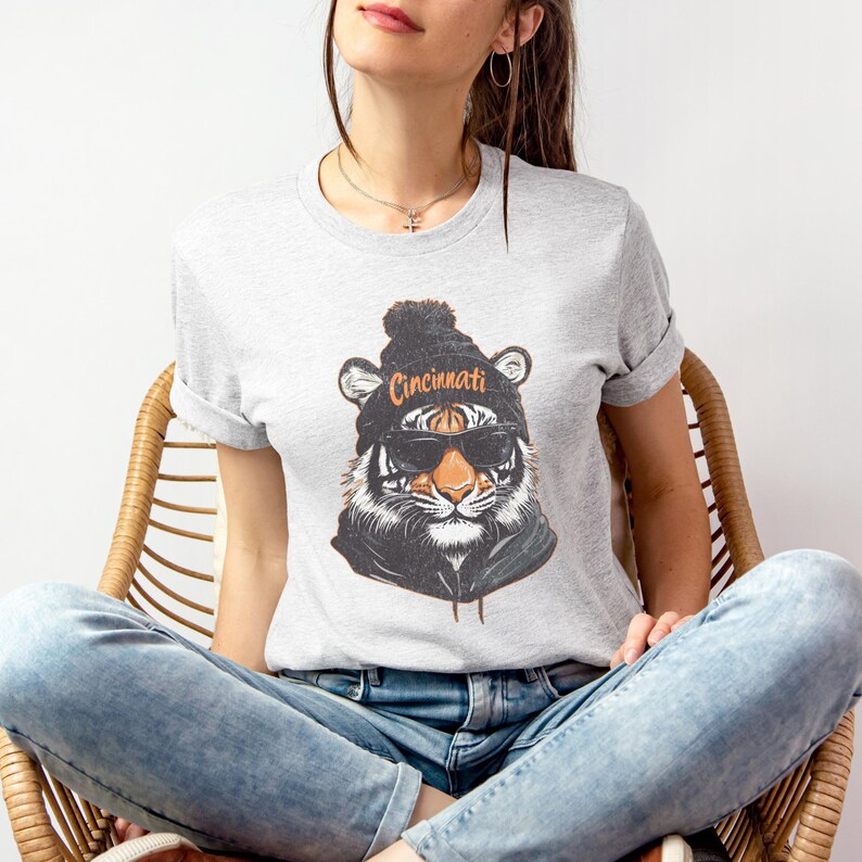 Puede incluir: Una camiseta gris con un gr&aacute;fico de un tigre que lleva una boina negra con la palabra "Cincinnati" en ella. El tigre lleva gafas de sol y tiene un patr&oacute;n de rayas blancas y negras.