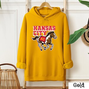 May include: A gold-colored hoodie with a front pocket and drawstring hood. The front features the text "KANSAS CITY" above a graphic of a horse with "KC" on its saddle. The hoodie is hanging on a wooden hanger.