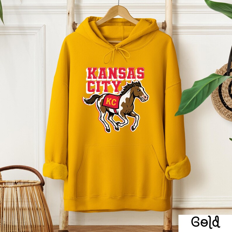 May include: A gold-colored hoodie with a front pocket and drawstring hood. The front features the text "KANSAS CITY" above a graphic of a horse with "KC" on its saddle. The hoodie is hanging on a wooden hanger.