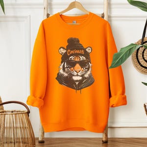Puede incluir: Sudadera naranja con un gr&aacute;fico de un tigre con una boina negra con la palabra "Cincinnati" y gafas de sol.