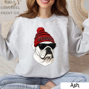 May include: Ash gray sweatshirt featuring a bulldog wearing a red and black beanie with the word "GEORGIA" and black sunglasses. The bulldog graphic is a detailed illustration. The sweatshirt is a casual style.