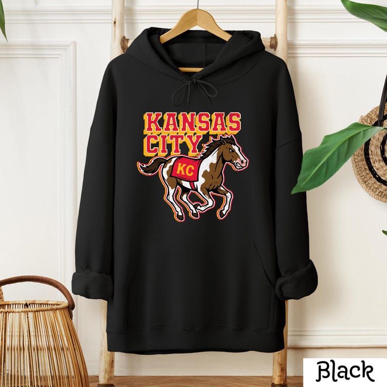 May include: Black hooded sweatshirt featuring a graphic of a horse and the words "Kansas City" in yellow and red. The horse has brown and white markings and a red "KC" on its side. The word "Black" is printed at the bottom.