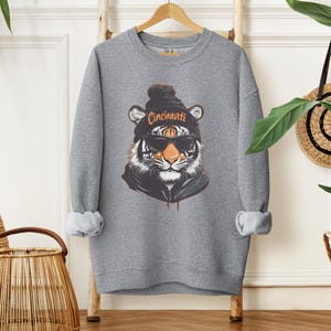Puede incluir: Sudadera gris con un gr&aacute;fico de un tigre con una boina con la palabra "Cincinnati" en ella. El tigre lleva gafas de sol y una sudadera con capucha negra.