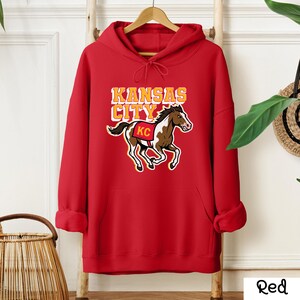 May include: A red hooded sweatshirt with a graphic of a horse and the words "Kansas City" in yellow and white. The horse has brown and white markings and a red saddle with "KC" on it.