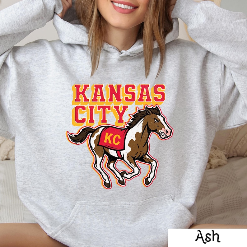 May include: Light grey hoodie with the text "KANSAS CITY" in yellow and red. A brown and white horse graphic with a red saddle displaying "KC" is featured below the text. The hoodie is made of a soft material.