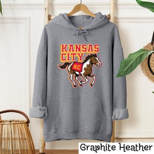 May include: A graphite heather gray hoodie with a graphic of a running horse and the words "KANSAS CITY" in yellow and red. The horse has a red and white saddle with "KC" on it. The text "Graphite Heather" is at the bottom.