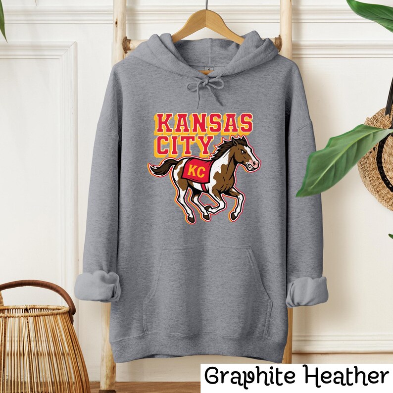 May include: A graphite heather gray hoodie with a graphic of a running horse and the words "KANSAS CITY" in yellow and red. The horse has a red and white saddle with "KC" on it. The text "Graphite Heather" is at the bottom.