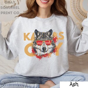 May include: A gray sweatshirt with a graphic print of a wolf wearing sunglasses and the text "KANSAS CITY" in gold lettering. The wolf is surrounded by red and orange paint splatters.