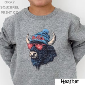 May include: A heather gray sweatshirt featuring a graphic of a buffalo wearing a blue beanie with "Buffalo" in red text, and red sunglasses. The sweatshirt has long sleeves and a crew neck. The text "GRAY SQUIRREL PRINT CO" is printed on the upper left.