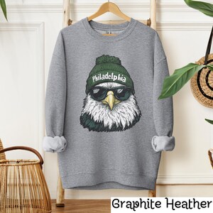 Op de afbeelding: Een grijs sweatshirt met een afbeelding van een Amerikaanse zeearend die een groene beanie draagt met het woord "Philadelphia" erop. De arend draagt een zonnebril.