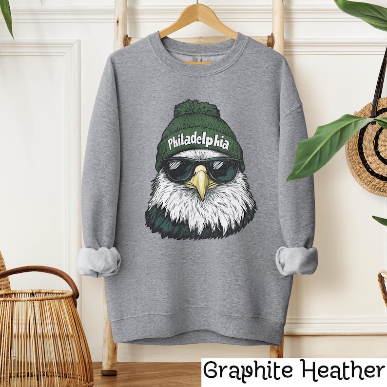 Op de afbeelding: Een grijs sweatshirt met een afbeelding van een Amerikaanse zeearend die een groene beanie draagt met het woord "Philadelphia" erop. De arend draagt een zonnebril.