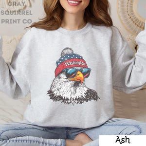 May include: Gray sweatshirt with a graphic of an eagle wearing a red, white, and blue beanie with the word "Washington" on it. The eagle is wearing sunglasses and has a serious expression.