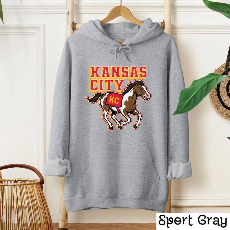May include: Sport gray hoodie featuring a graphic of a running horse with "Kansas City" in red and yellow text. The horse has brown and white markings and a red "KC" logo. The hoodie has a drawstring hood and a kangaroo pocket.
