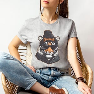 Puede incluir: Una camiseta gris con un gr&aacute;fico de un tigre que lleva una boina negra con la palabra "Cincinnati" en ella. El tigre lleva gafas de sol.