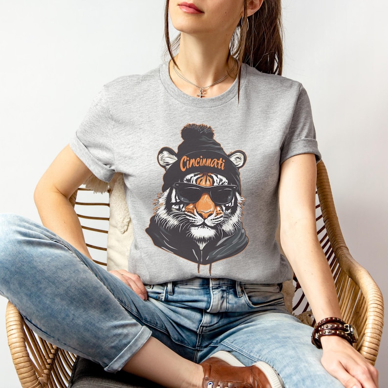 Puede incluir: Una camiseta gris con un gr&aacute;fico de un tigre que lleva una boina negra con la palabra "Cincinnati" en ella. El tigre lleva gafas de sol.