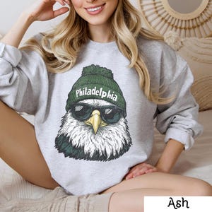 May include: A gray sweatshirt with a graphic of a bald eagle wearing a green hat that says "Philadelphia" and sunglasses. The sweatshirt is available in the color Ash.