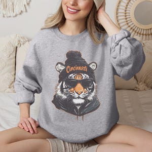 Puede incluir: Sudadera gris con un gr&aacute;fico de un tigre que lleva una boina negra con un pomp&oacute;n blanco y gafas de sol. La boina tiene la palabra "Cincinnati" escrita en ella.