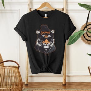 Puede incluir: Camiseta negra con un gr&aacute;fico de un tigre con una boina con la palabra "Cincinnati" en ella. El tigre lleva gafas de sol.