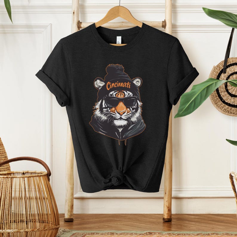 Puede incluir: Camiseta negra con un gr&aacute;fico de un tigre con una boina con la palabra "Cincinnati" en ella. El tigre lleva gafas de sol.
