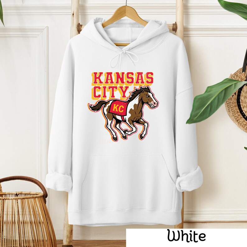 May include: White hoodie with a kangaroo pocket and drawstring hood. The front features the words "Kansas City" in yellow and orange, with a brown and white horse graphic and the letters "KC".