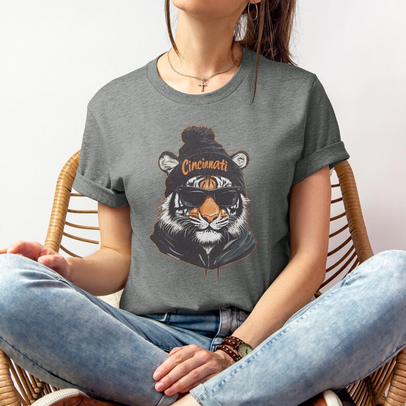 Puede incluir: Una camiseta gris con un gr&aacute;fico de un tigre que lleva una boina negra con la palabra "Cincinnati" en ella. El tigre lleva gafas de sol y una sudadera con capucha negra.
