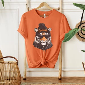 Puede incluir: Camiseta naranja con un gr&aacute;fico de un tigre con una boina negra con la palabra "Cincinnati" y gafas de sol.