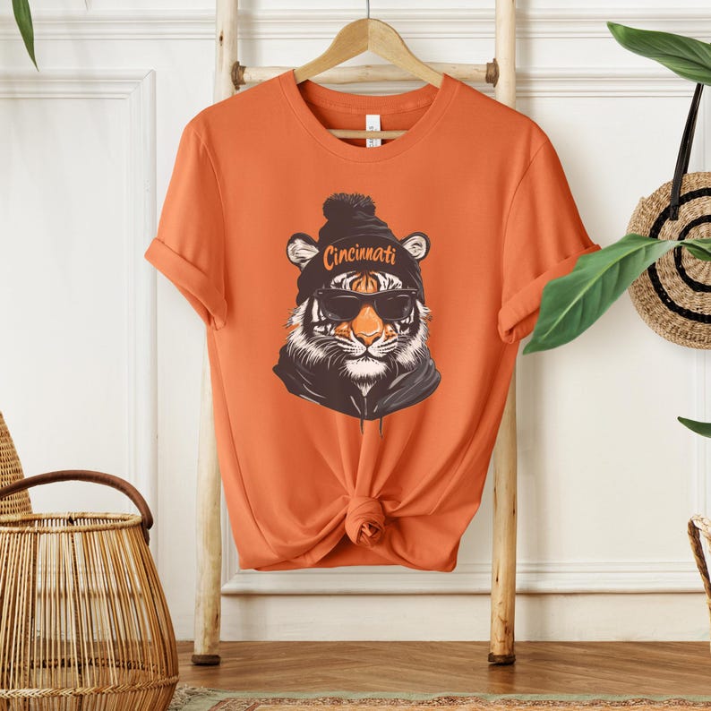 Puede incluir: Camiseta naranja con un gr&aacute;fico de un tigre con una boina negra con la palabra "Cincinnati" y gafas de sol.