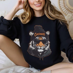 Puede incluir: Sudadera negra con un gr&aacute;fico de un tigre que lleva una boina negra con un pomp&oacute;n blanco y gafas de sol. La boina tiene la palabra "Cincinnati" escrita en naranja.