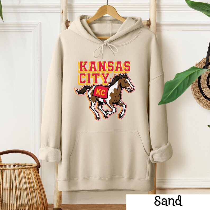May include: A sand-colored hoodie with a kangaroo pocket and drawstring hood. The front features the text "KANSAS CITY" in yellow and orange, with a brown and white horse graphic and "KC" on its saddle.