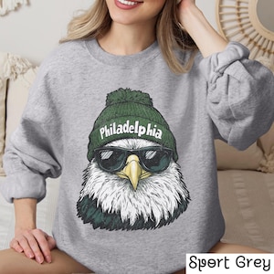 Op de afbeelding: Een grijs sweatshirt met een afbeelding van een Amerikaanse zeearend die een groene beanie draagt met het woord "Philadelphia" erop. De adelaar draagt een zonnebril.