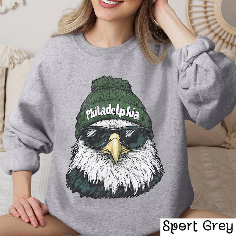 Op de afbeelding: Een grijs sweatshirt met een afbeelding van een Amerikaanse zeearend die een groene beanie draagt met het woord "Philadelphia" erop. De adelaar draagt een zonnebril.