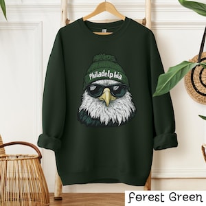 Op de afbeelding: Bosgroen sweatshirt met een afbeelding van een Amerikaanse zeearend die een groene beanie draagt met het woord "Philadelphia" erop en een zonnebril.