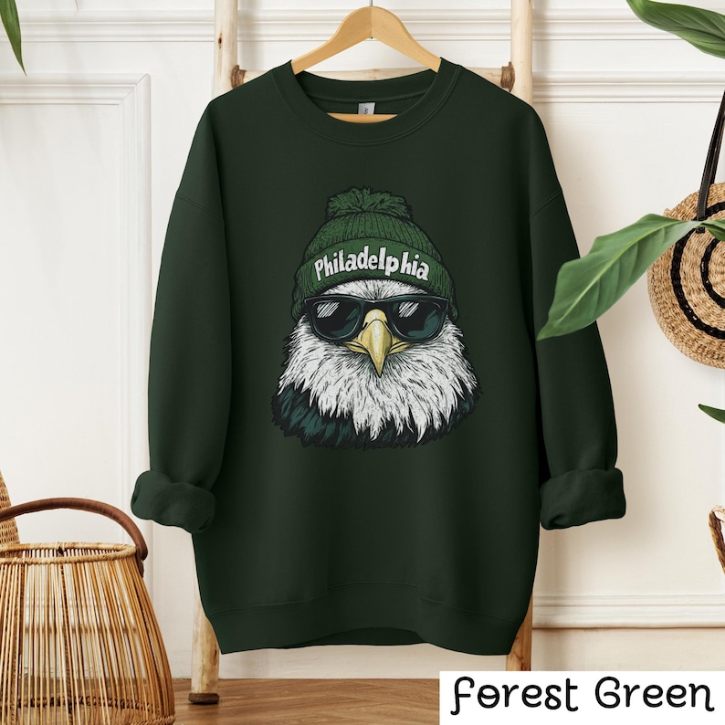 Op de afbeelding: Bosgroen sweatshirt met een afbeelding van een Amerikaanse zeearend die een groene beanie draagt met het woord "Philadelphia" erop en een zonnebril.