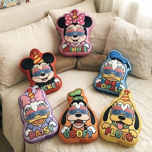 May include: A collection of character-shaped throw pillows featuring Mickey Mouse, Minnie Mouse, Donald Duck, Daisy Duck, Goofy, and Pluto. Each pillow has sunglasses and a name. The pillows are in various colors, including red, pink, blue, purple, orange, and yellow.