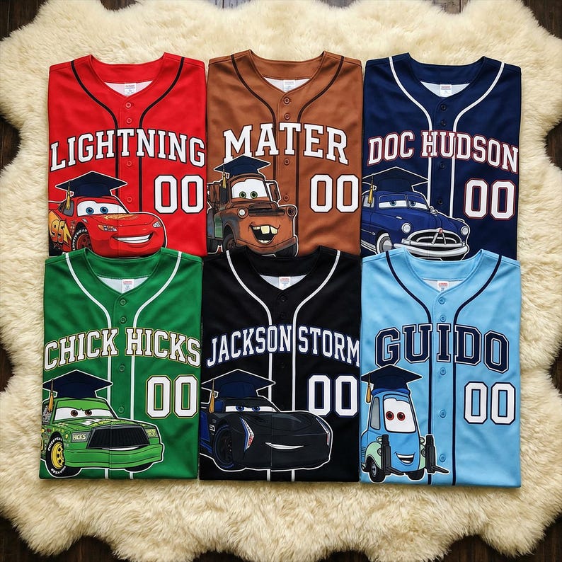 May include: Six baseball jerseys in red, brown, navy, green, black, and light blue. Each jersey features a cartoon car character, name, and the number 00. The jerseys are laid out on a white, fluffy surface.